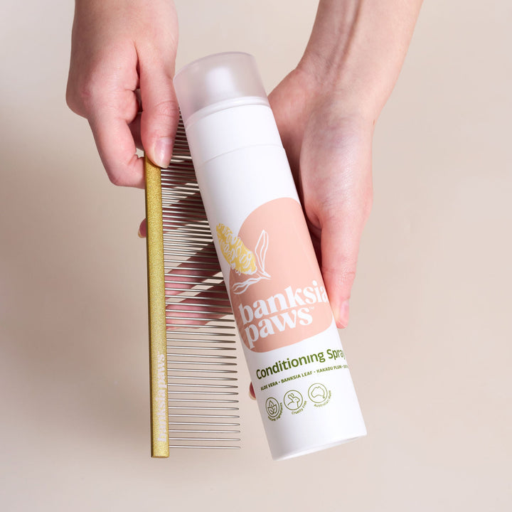Person holding a bottle of 'Banksia Paws' conditioning spray next to a comb on a light background